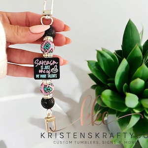 May include: A beaded keychain with a gold-colored clasp. The keychain features black, pink, and turquoise beads, and a black square with the text "Sarcasm is just one of my many talents".
