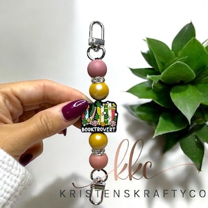May include: A beaded keychain with a silver clasp and a charm that reads "BOOKTROVERT." The keychain features alternating pink and yellow beads, with rhinestone accents. The charm has a colorful design of books and flowers.