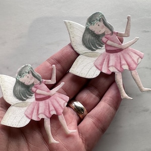 K&ouml;nnte beinhalten: Zwei Papierschablonen von Feen. Jede Fee hat graues Haar, einen Blumenkranz und ein rosa Kleid. Sie haben wei&szlig;e und beige Fl&uuml;gel. Die Feen befinden sich in einer Tanzpose mit erhobenen Armen. Das Bild ist eine Nahaufnahme.
