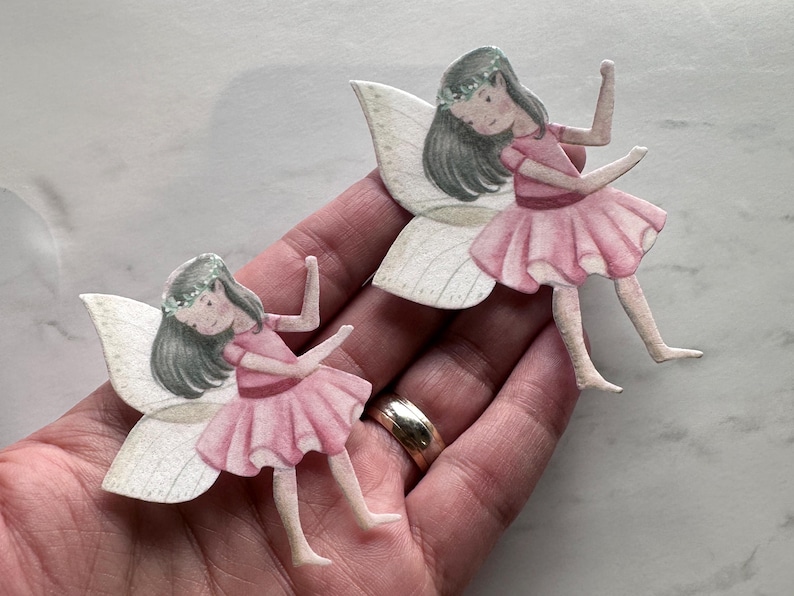 K&ouml;nnte beinhalten: Zwei Papierschablonen von Feen. Jede Fee hat graues Haar, einen Blumenkranz und ein rosa Kleid. Sie haben wei&szlig;e und beige Fl&uuml;gel. Die Feen befinden sich in einer Tanzpose mit erhobenen Armen. Das Bild ist eine Nahaufnahme.