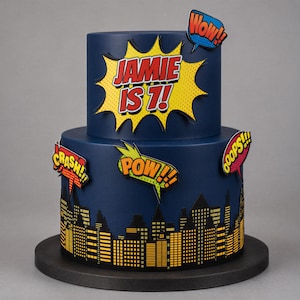 Può includere: Una torta a due piani blu scuro con un'esplosione in stile fumetto gialla con il testo JAMIE IS 7!. Il piano inferiore mostra una silhouette di città nera con finestre gialle. I fumetti riportano le scritte WOW!, CRASH!, POW! e OOPS!.