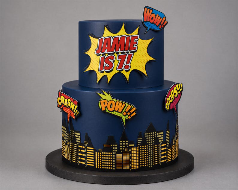 Puede incluir: Un pastel de dos niveles azul oscuro con una explosi&oacute;n estilo c&oacute;mic amarilla con el texto JAMIE IS 7!. El nivel inferior muestra una silueta de ciudad negra con ventanas amarillas. Los globos de texto de c&oacute;mic dicen WOW!, CRASH!, POW! y OOPS!.