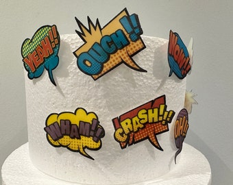 May include: A white cake decorated with colorful comic book speech bubbles with words like "Yeah!" "Ouch!" "Wow!" "Wham!" "Crash!" and "Oh!"