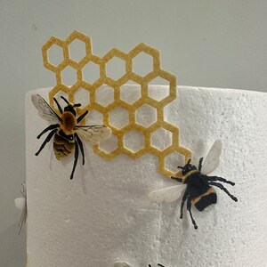 Pu&ograve; includere: Una base cilindrica in schiuma bianca decorata con un ritaglio in feltro giallo a forma di alveare e due figure di api realistiche. Le api presentano corpi a strisce gialle e nere, zampe scure e ali traslucide fissate sulla schiuma bianca.
