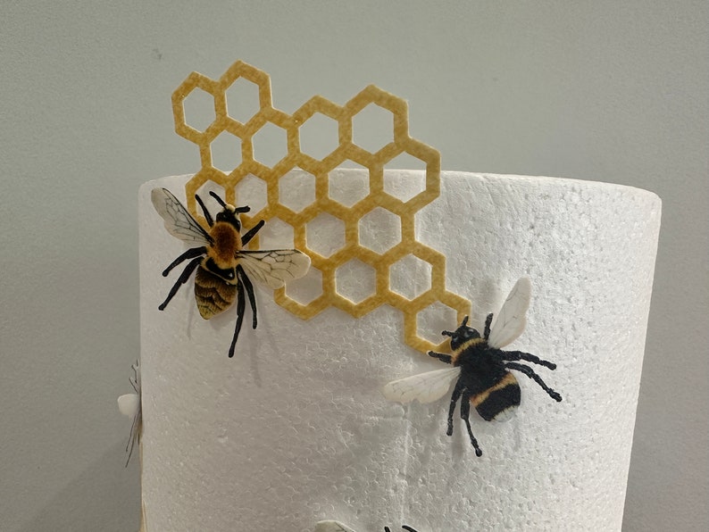 Pu&ograve; includere: Una base cilindrica in schiuma bianca decorata con un ritaglio in feltro giallo a forma di alveare e due figure di api realistiche. Le api presentano corpi a strisce gialle e nere, zampe scure e ali traslucide fissate sulla schiuma bianca.