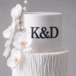 May include: A two-tiered white cake decorated with white orchids and the letters "K&D" in navy blue. The top tier is smooth, while the bottom tier has a textured, ruffled design. The cake is a wedding or celebration cake.