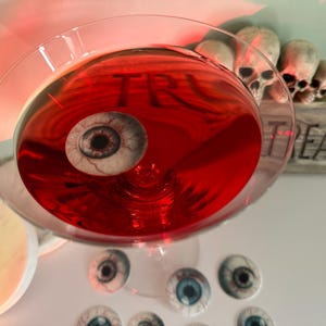 Puede incluir: Un c&oacute;ctel rojo en una copa de martini con un ojo falso flotando en &eacute;l. La copa est&aacute; rodeada de varios ojos falsos m&aacute;s. Un letrero de madera con la palabra "Treat" est&aacute; en el fondo.