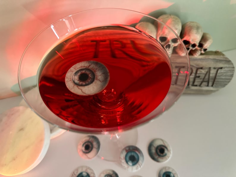 Puede incluir: Un c&oacute;ctel rojo en una copa de martini con un ojo falso flotando en &eacute;l. La copa est&aacute; rodeada de varios ojos falsos m&aacute;s. Un letrero de madera con la palabra "Treat" est&aacute; en el fondo.