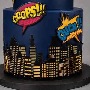 Puede incluir: Un pastel cil&iacute;ndrico azul oscuro decorado con una silueta negra de un horizonte urbano con ventanas amarillas. Dos globos de texto estilo c&oacute;mic est&aacute;n adheridos al lado, uno rosa con el texto OOOPS!!! y uno amarillo con OUCH!!