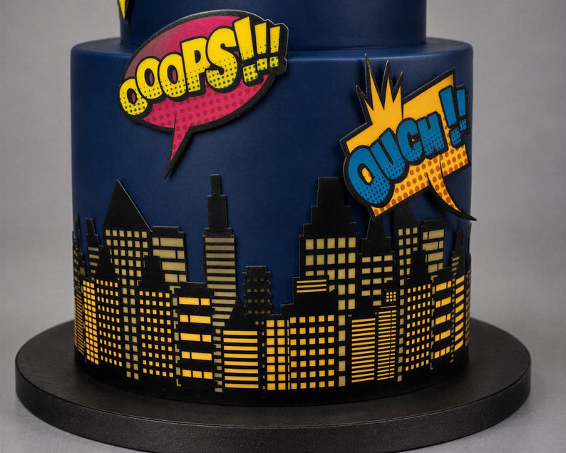 Puede incluir: Un pastel cil&iacute;ndrico azul oscuro decorado con una silueta negra de un horizonte urbano con ventanas amarillas. Dos globos de texto estilo c&oacute;mic est&aacute;n adheridos al lado, uno rosa con el texto OOOPS!!! y uno amarillo con OUCH!!