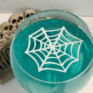 May include: A glass filled with a teal blue liquid and a white spider web design on top. The glass is sitting on a wooden surface with the word "Trick" written on it.