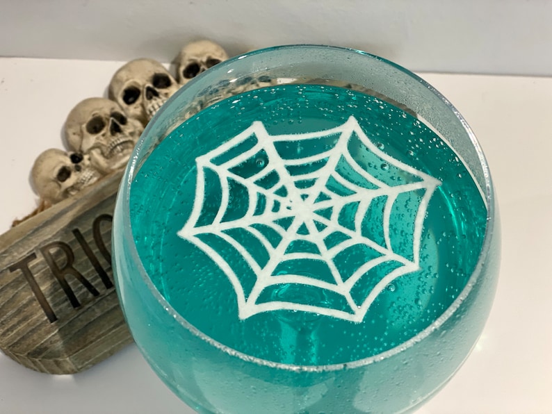 May include: A glass filled with a teal blue liquid and a white spider web design on top. The glass is sitting on a wooden surface with the word "Trick" written on it.