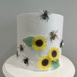 Pu&ograve; includere: Un manichino per torta cilindrico in schiuma bianca decorato con tre girasoli gialli, due foglie verdi e cinque piccole figure di calabroni neri e gialli fissate sulla superficie esterna. L'oggetto &egrave; posizionato su un'alzata bianca.