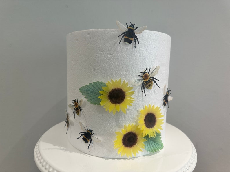 Pu&ograve; includere: Un manichino per torta cilindrico in schiuma bianca decorato con tre girasoli gialli, due foglie verdi e cinque piccole figure di calabroni neri e gialli fissate sulla superficie esterna. L'oggetto &egrave; posizionato su un'alzata bianca.