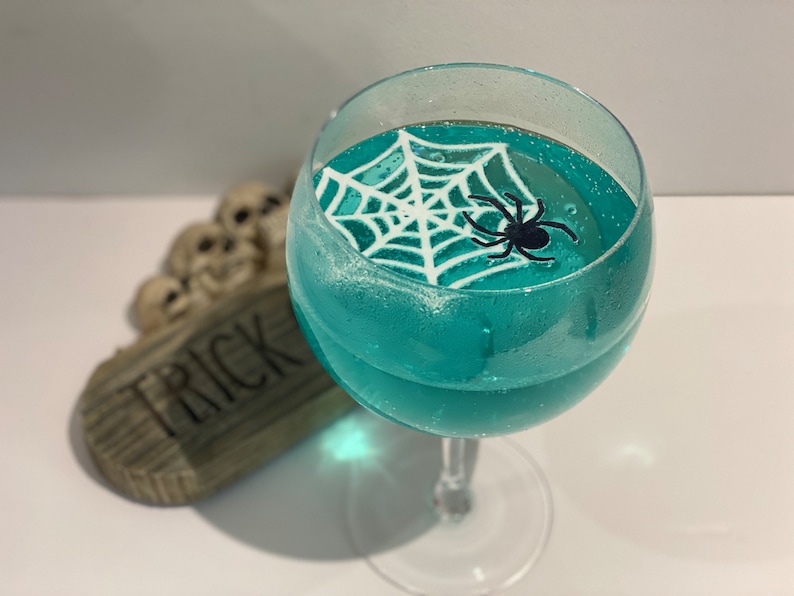 May include: A teal blue cocktail in a stemmed glass with a white spiderweb and a black spider on top. The glass is sitting on a wooden surface with the word "TRICK" written on it.