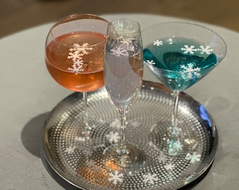 May include: Three stemmed glasses with colorful drinks, including a rose-colored wine, a clear champagne, and a teal cocktail, each adorned with snowflake decorations. The glasses sit on a silver tray.