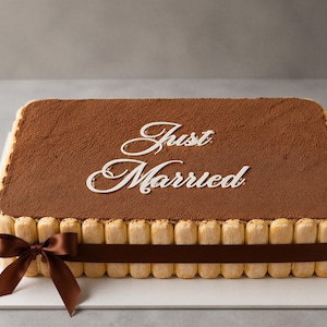 May include: A rectangular tiramisu cake topped with cocoa powder and white script text that reads Just Married. The sides are lined with ladyfinger biscuits, and a dark brown satin ribbon is tied in a bow around the perimeter of the cake.