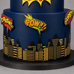Puede incluir: Un pastel azul oscuro de varios niveles decorado con una silueta de ciudad negra y amarilla en la base. El pastel presenta burbujas de di&aacute;logo estilo c&oacute;mic con el texto CRASH, POW y OOPS en colores vibrantes amarillo, naranja y rojo.