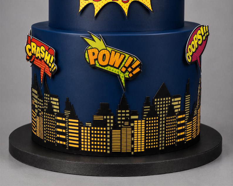 Puede incluir: Un pastel azul oscuro de varios niveles decorado con una silueta de ciudad negra y amarilla en la base. El pastel presenta burbujas de di&aacute;logo estilo c&oacute;mic con el texto CRASH, POW y OOPS en colores vibrantes amarillo, naranja y rojo.