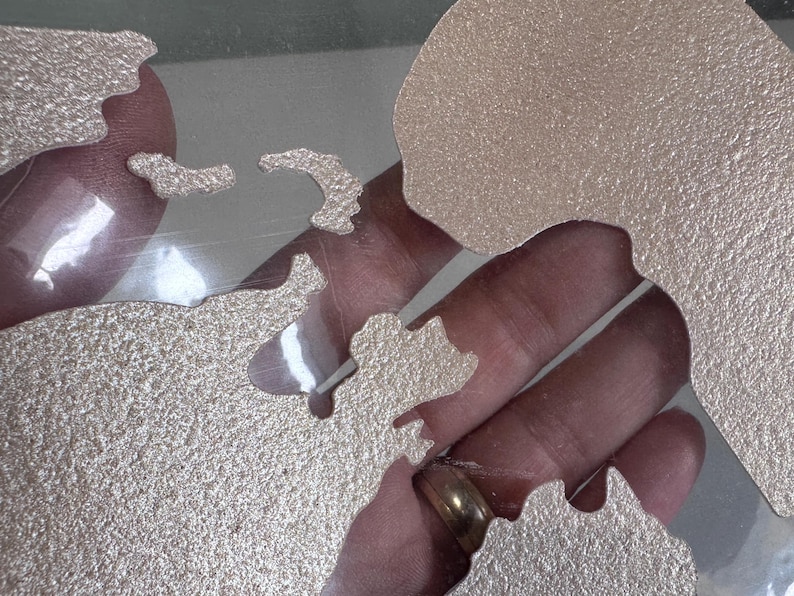 May include: A set of metallic gold-colored stencils in the shape of continents. The stencils are on a clear surface.