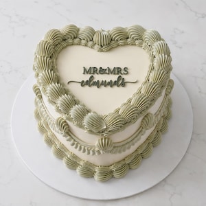 Könnte beinhalten: Eine herzförmige Torte mit glattem cremefarbenem Zuckerguss, verziert mit salbeigrünen Buttercreme-Rändern und Girlanden. Ein dunkelgrauer Acryl-Tortentopper ist mittig platziert und trägt den Text MR & MRS edmunds in verschiedenen Schriftarten.