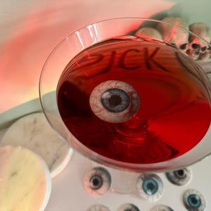 Puede incluir: Un c&oacute;ctel rojo en una copa de martini con un ojo falso flotando en &eacute;l. La copa est&aacute; sobre una superficie blanca con varios ojos falsos esparcidos a su alrededor.