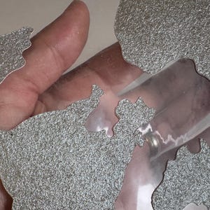 May include: A close-up of a silver glitter-covered world map cut out of paper. The map is placed on a clear plastic sheet.