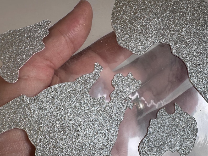 May include: A close-up of a silver glitter-covered world map cut out of paper. The map is placed on a clear plastic sheet.