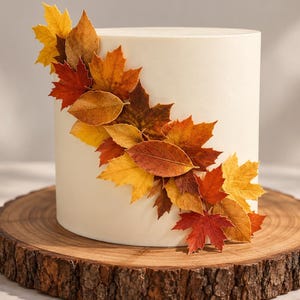 May include: A cylindrical cake with smooth white frosting decorated with a diagonal arrangement of edible autumn leaves in shades of yellow, orange, and deep red, resting on a circular wooden slab with visible bark edges.