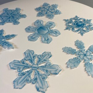 May include: Seven blue snowflake cutouts on a white background. The snowflakes are various sizes and shapes.