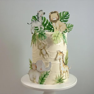May include: A cylindrical cake with white textured frosting decorated with paper cutouts of a zebra, lion, monkey, elephant, and giraffe, interspersed with green tropical palm leaves, all arranged on a white cake stand.