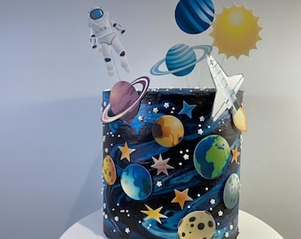 May include: A cylindrical cake with dark blue and black marbled frosting decorated with various planet cutouts, stars, and small white sprinkles. Cake toppers include an astronaut, a space shuttle, a yellow sun, and several planets on sticks.