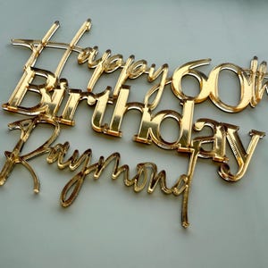 May include: A gold-colored cake topper with the words "Happy 60th Birthday Raymond" in a script font. The topper is made of a shiny material and is designed to be placed on top of a cake for a birthday celebration.