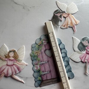 K&ouml;nnte beinhalten: Aquarellillustrationen von Feen und einer Feent&uuml;r. Die Feen tragen rosa und gelbe Kleider mit wei&szlig;en Fl&uuml;geln. Ein Holzlineal liegt neben der Feent&uuml;r, die etwa 10 cm hoch ist.