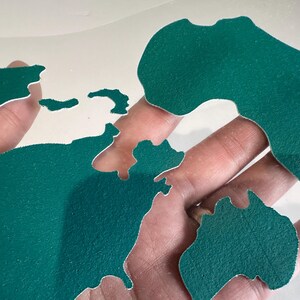 May include: A green world map cut out of felt, with each continent separated. The map is held in a hand.