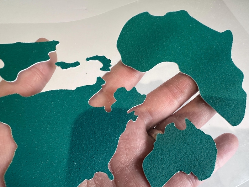 May include: A green world map cut out of felt, with each continent separated. The map is held in a hand.