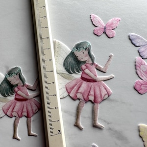 May include: Two pink and white fairy figurines with wings and floral headbands. The fairies are wearing pink dresses and have brown hair. The background is white with several pink, purple, and yellow butterflies.