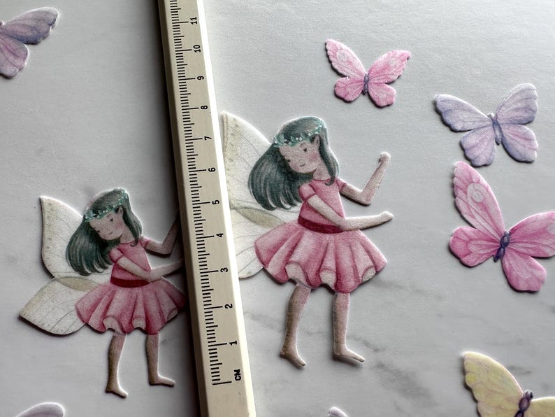 May include: Two pink and white fairy figurines with wings and floral headbands. The fairies are wearing pink dresses and have brown hair. The background is white with several pink, purple, and yellow butterflies.