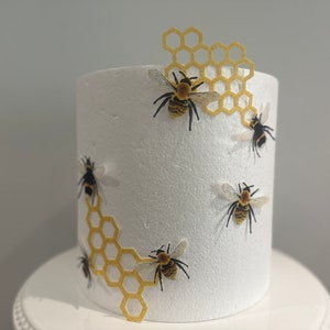 Pu&ograve; includere: Un cilindro in schiuma bianca per torte decorato con ritagli gialli a forma di nido d'ape e diverse piccole figure di api realistiche. Le api presentano corpi a strisce nere e gialle con ali traslucide sulla superficie bianca.