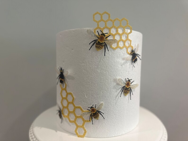 Pu&ograve; includere: Un cilindro in schiuma bianca per torte decorato con ritagli gialli a forma di nido d'ape e diverse piccole figure di api realistiche. Le api presentano corpi a strisce nere e gialle con ali traslucide sulla superficie bianca.