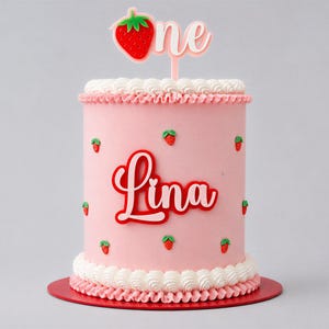 Peut inclure: Un gâteau rose décoré de glaçage blanc et d'accents rouges. Le gâteau est orné de petites fraises et du nom "Lina" en écriture rouge. Une fraise et le mot "one" sont sur le dessus.