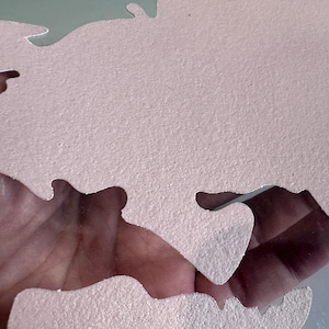 May include: A pink, textured, foam puzzle piece shaped like a map of the world. The piece is cut out in a way that shows a hand holding it.