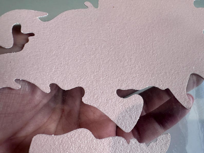 May include: A pink, textured, foam puzzle piece shaped like a map of the world. The piece is cut out in a way that shows a hand holding it.