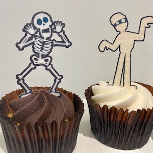 May include: Two Halloween cupcakes decorated with edible toppers. One cupcake has a chocolate frosting and a skeleton topper. The other cupcake has a white frosting and a mummy topper.