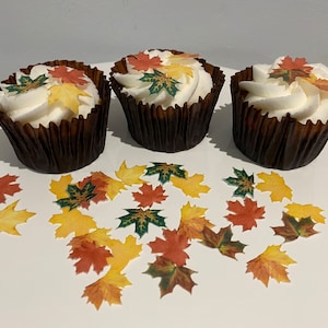 May include: Three cupcakes decorated with autumn-themed edible leaves. The cupcakes are topped with white icing and have brown paper cases. The leaves are in shades of red, orange, yellow, and green.