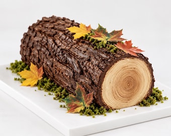Puede incluir: Un pastel de tronco de Navidad de chocolate con forma de tronco de árbol, corteza marrón oscuro texturizada, sección transversal color canela con anillos de madera, musgo comestible verde y hojas de arce otoñales de colores en la base.