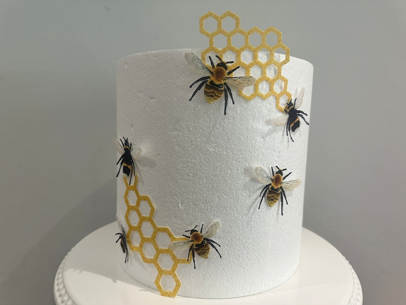 Pu&ograve; includere: Un manichino per torta cilindrico in schiuma bianca decorato con ritagli a nido d'ape gialli e diverse figure di api realistiche. Le api presentano corpi a strisce nere e gialle con ali traslucide sulla superficie bianca testurizzata.