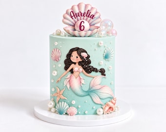 Puede incluir: Un pastel cilíndrico de color azul claro decorado con una ilustración de sirena, detalles de perlas blancas y varias conchas marinas. Un adorno de concha rosa grande muestra el nombre Aurelia y el número 6 sobre una perla.