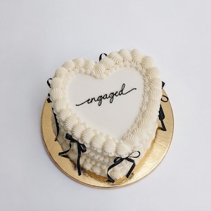 May include: A heart-shaped cake with smooth white icing, featuring the word engaged written in black script on top. The cake is decorated with white piped icing borders and small black ribbon bows attached to the sides, resting on a gold base.