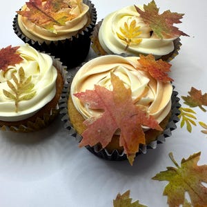 May include: Four cupcakes decorated with autumn-themed edible leaves. The cupcakes have white and peach-coloured frosting. The leaves are in shades of orange, yellow, and green.